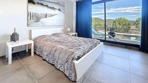 A bedroom with a bed, a nightstand, a wall-mounted air conditioner, a painting, and a large window with a view of a pool and landscape.
