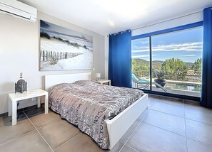 Quarto com cama, mesa de cabeceira, ar-condicionado na parede, pintura e uma grande janela com vista para uma piscina e paisagem.