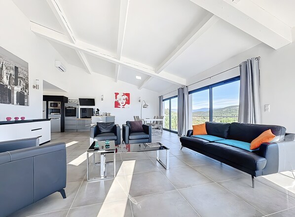 Uma sala de estar moderna com um sofá, cadeiras, uma mesa de centro e uma cozinha ao fundo.