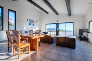 Un vaste espace de vie comprenant une table à manger en bois, des sièges confortables, un téléviseur à écran plat et de grandes fenêtres offrant une vue sur l’extérieur.