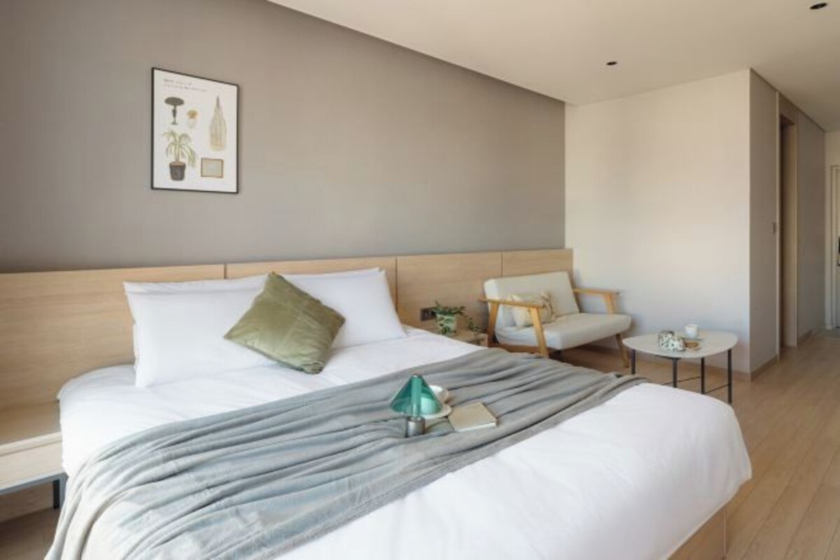 A modern hotel room with a large bed, a small side table, a chair, and a framed picture on the wall.