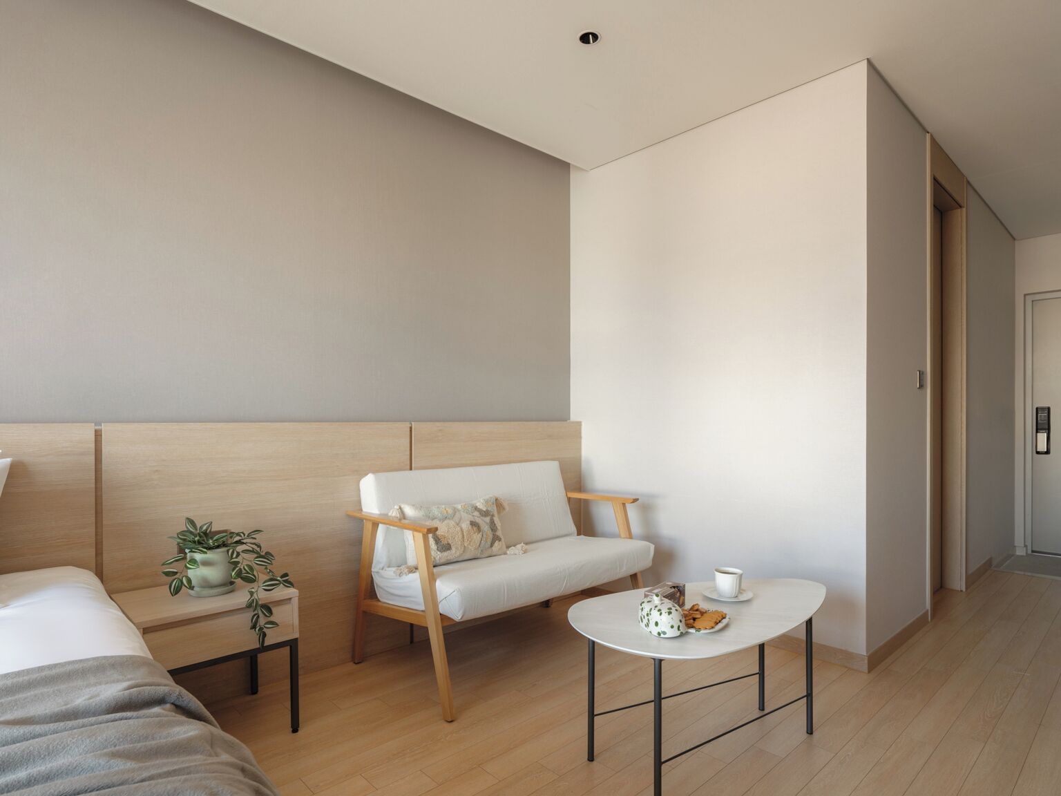 A modern hotel room with a bed, a sofa, a small table with a cup and cookies, and a plant on a nightstand.