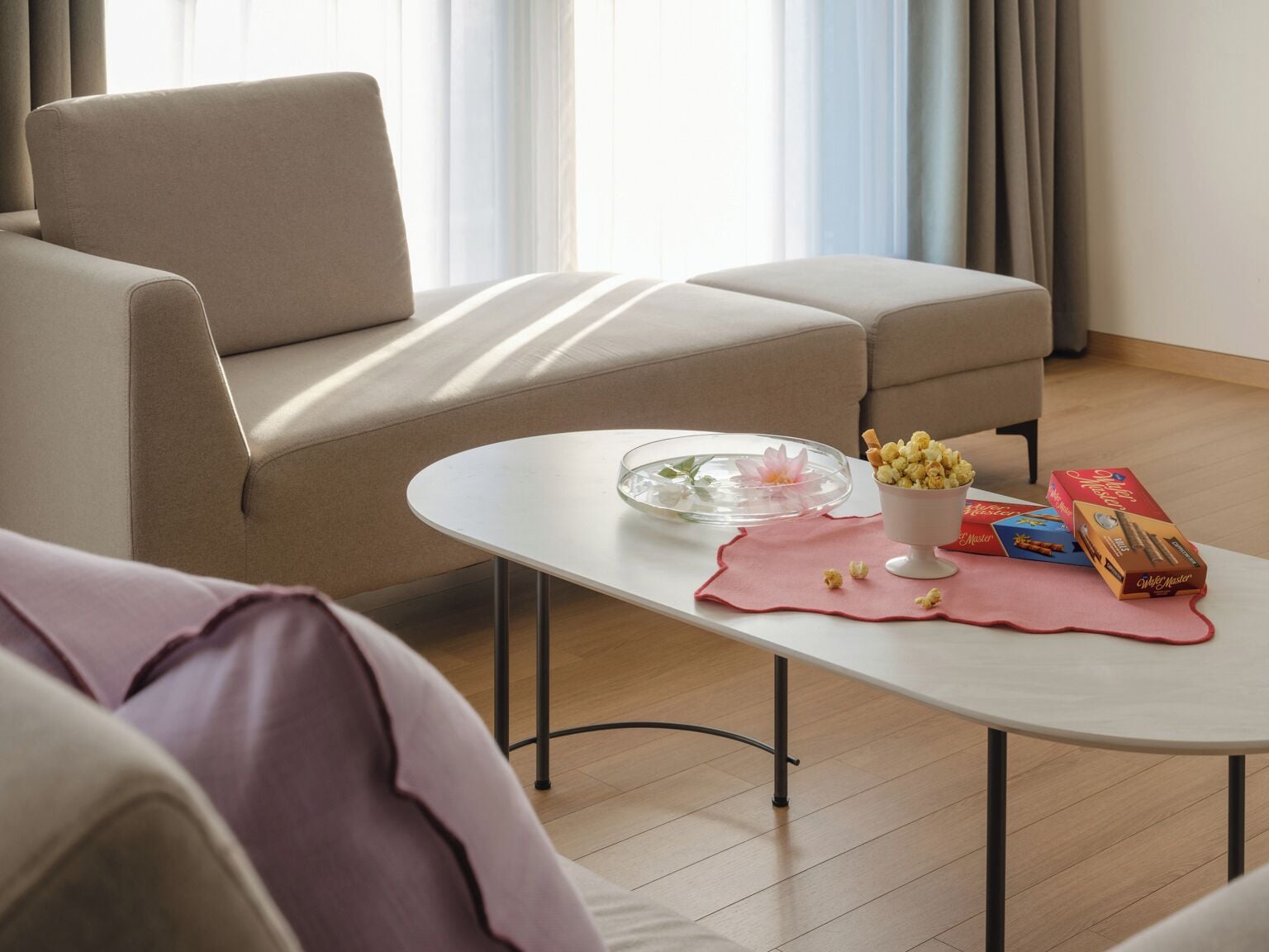 A modern living room with a sofa, a coffee table with snacks and a flower, and a view of the outdoors through large windows.