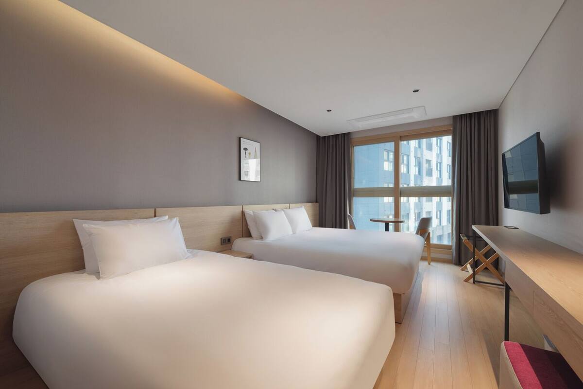 A hotel room with two beds, a desk, a chair, a TV, and a window with a city view.