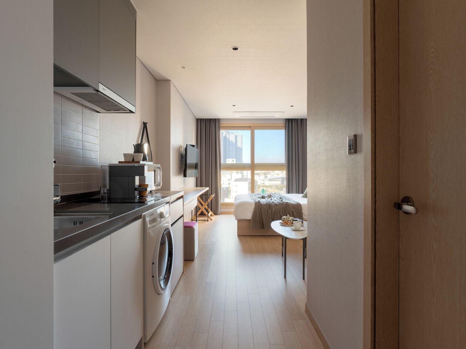 Quarto de hotel moderno com cozinha compacta, máquina de lavar, área de jantar e uma grande janela com vista para a cidade.