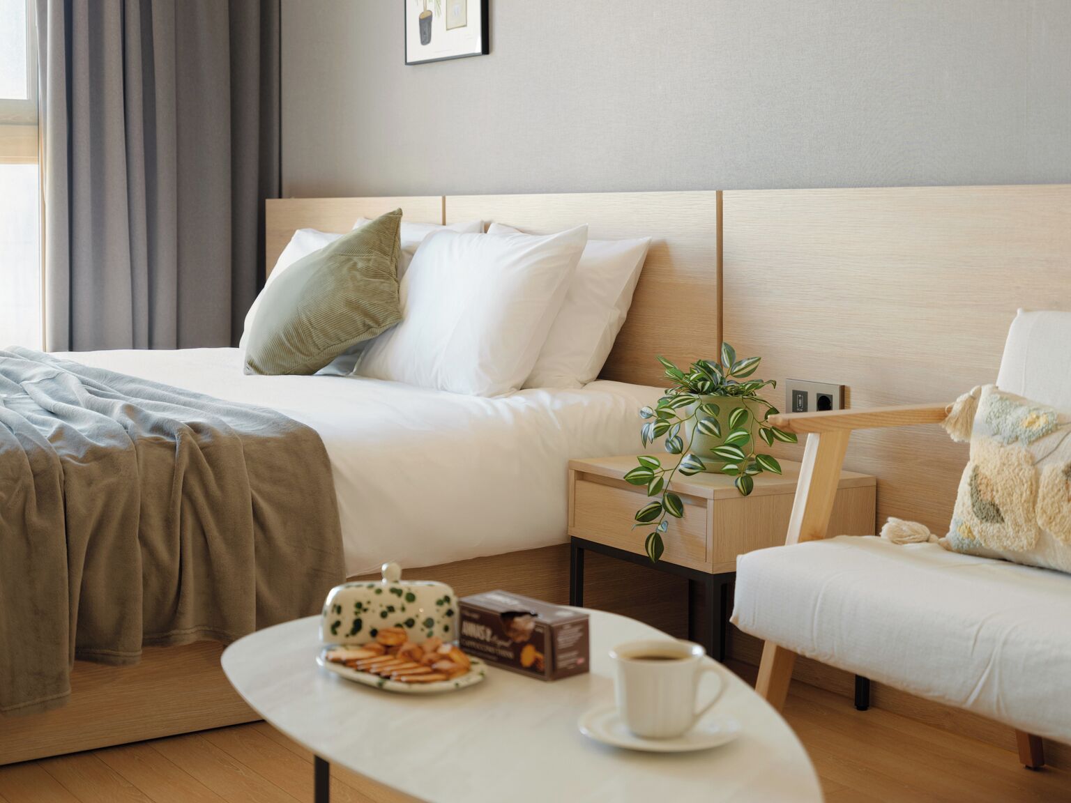 Habitación de hotel con cama, mesita de noche, silla y una pequeña mesa con aperitivos y una taza.