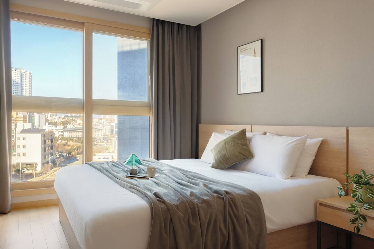 A hotel room with a large window, a bed with white linens, a green throw blanket, and a nightstand with a plant.
