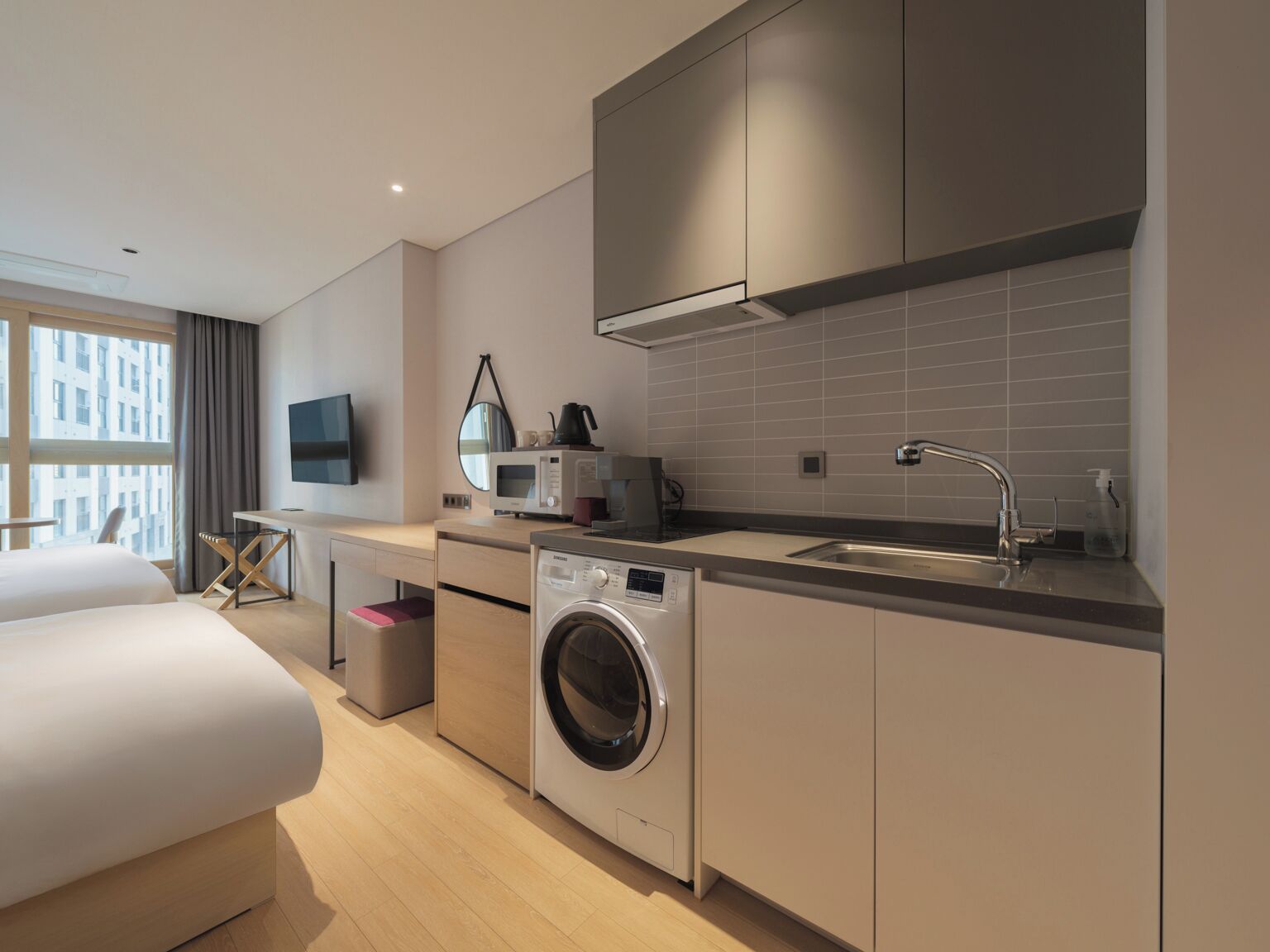 Una habitación de hotel moderna con una cama, lavadora, kitchenette y un gran ventanal.