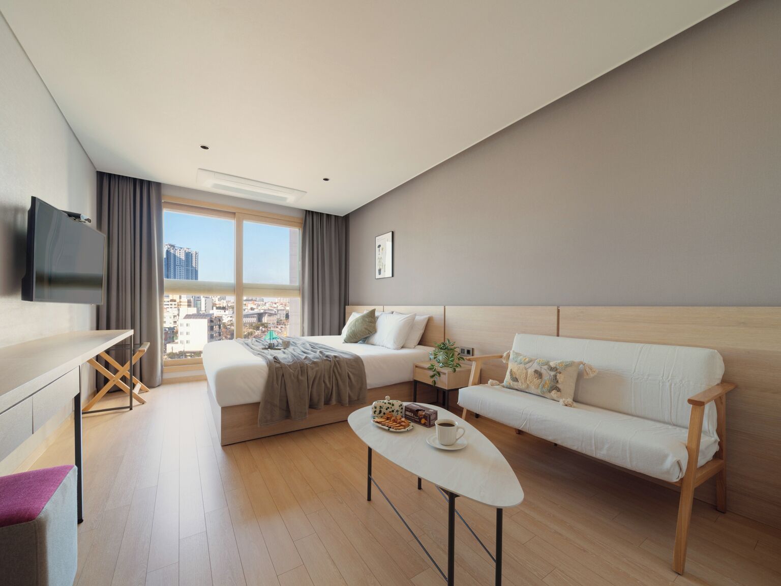 A modern hotel room with a bed, a sofa, a small table with snacks, a TV, and a city view.