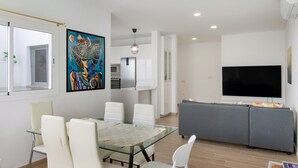 A modern living room with a glass dining table, white chairs, a gray sofa, a flat-screen TV, and a kitchen area with white cabinets.