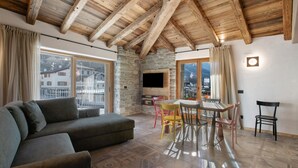 Un salon avec un mur en pierre, des poutres apparentes en bois au plafond, un téléviseur à écran plat, un canapé, une table à manger avec des chaises et de grandes fenêtres.
