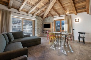 Ein Wohnzimmer mit Steinwand, Holzbalkendecke, Fernseher, Sofa, Esstisch mit Stühlen und großen Fenstern.