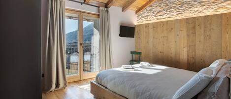 A bedroom with a wooden bed, a green chair, a flat-screen TV, and a view of mountains through a glass door.