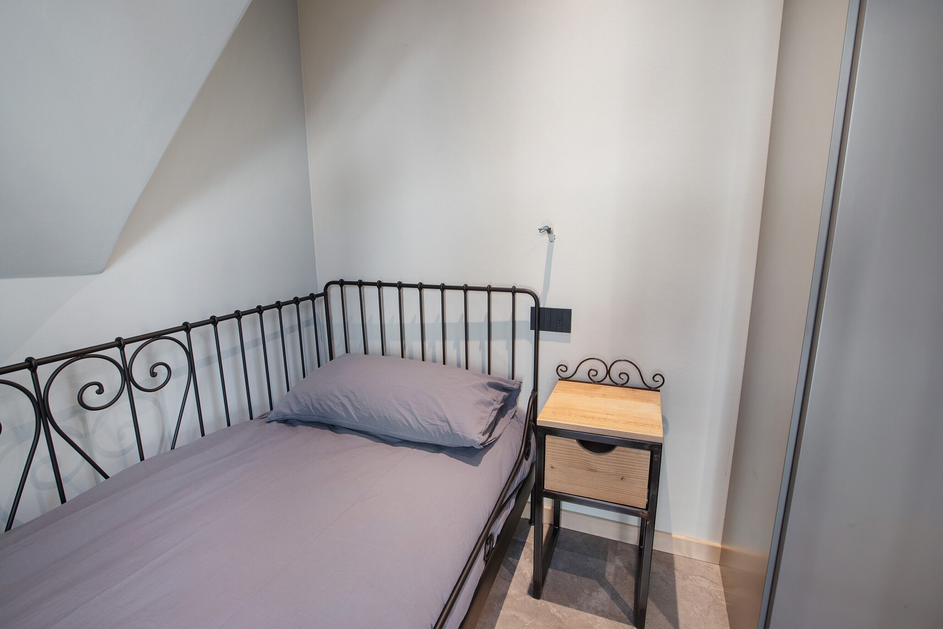 Un lit simple avec un cadre en métal, une table de chevet en bois avec un tiroir et un luminaire fixé au mur.