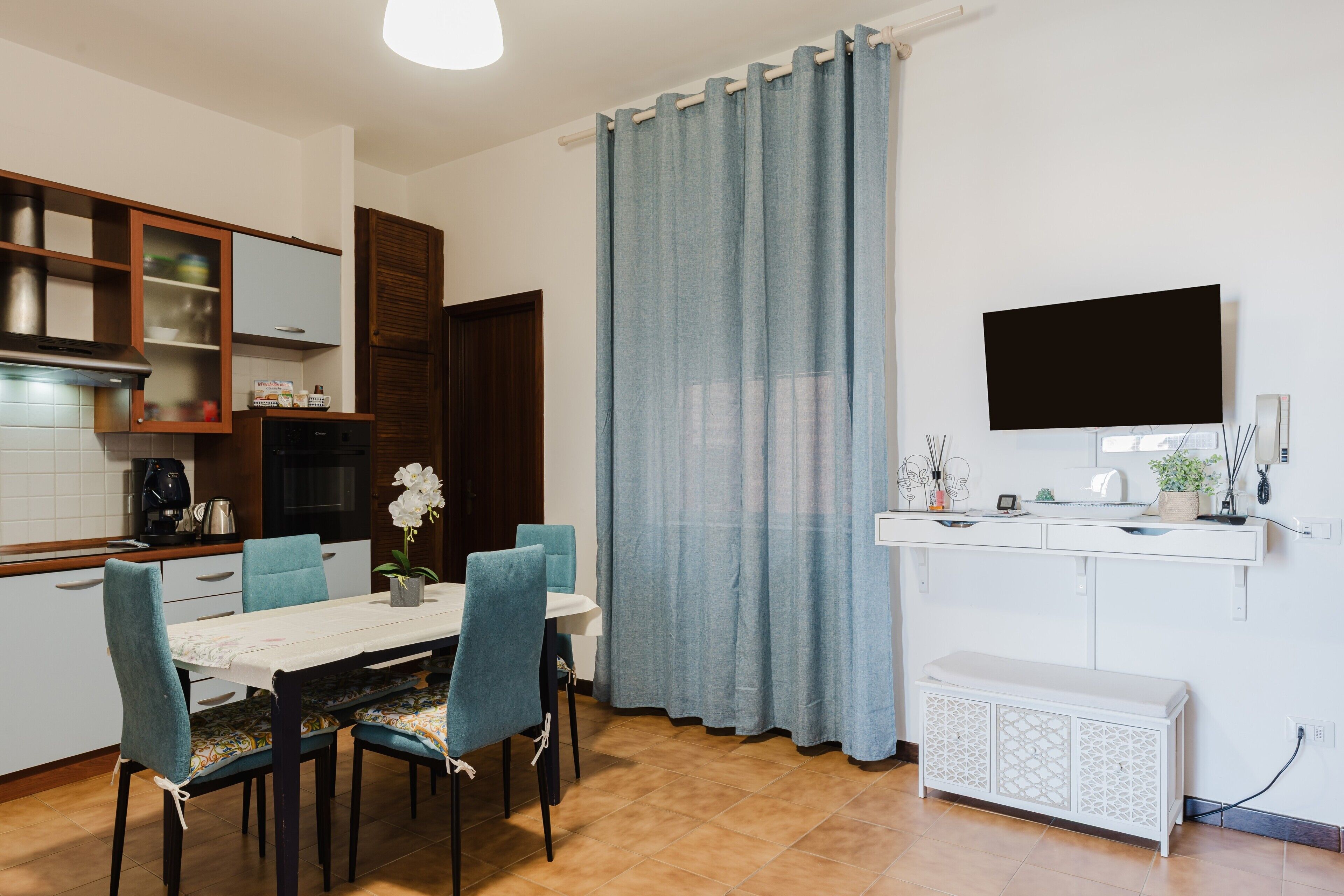 Een moderne keuken met een eethoek, een aan de muur bevestigde televisie en een groot raam met gordijnen.