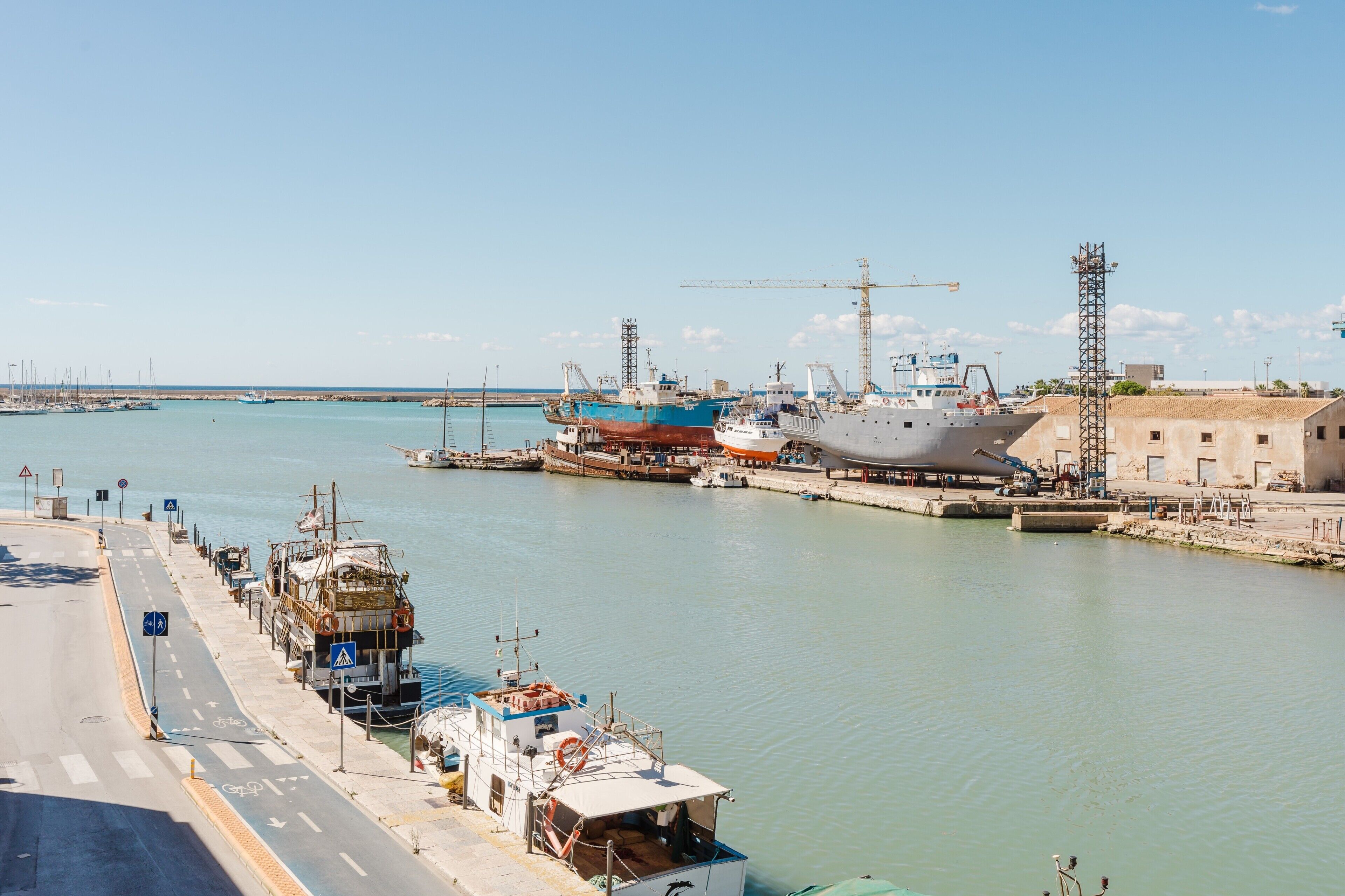 A marina with boats docked, a road parallel to the water, and industrial structures in the background.