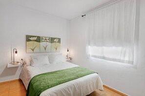 Une chambre à coucher avec un lit, un plaid vert, un tableau accroché au mur, des lampes de chevet et une fenêtre ornée de rideaux légers.