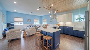 A spacious living area with a wooden ceiling, a kitchen with blue cabinets, and a large island with stools. - Luxe Coastal Stay w/ Private Pool, Walk to Beach | Leave of Absence by AvantStay (Port Aransas)