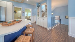 A kitchen with a white island, wooden chairs, and a staircase leading to a loft area. - Luxe Coastal Stay w/ Private Pool, Walk to Beach | Leave of Absence by AvantStay (Port Aransas)