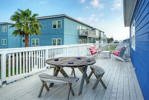 Une terrasse en bois avec une table ronde et des bancs, un palmier et un bâtiment bleu avec des balustrades blanches.