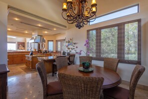A spacious kitchen with a central island, a dining area with wicker chairs, and large windows with a view of greenery.