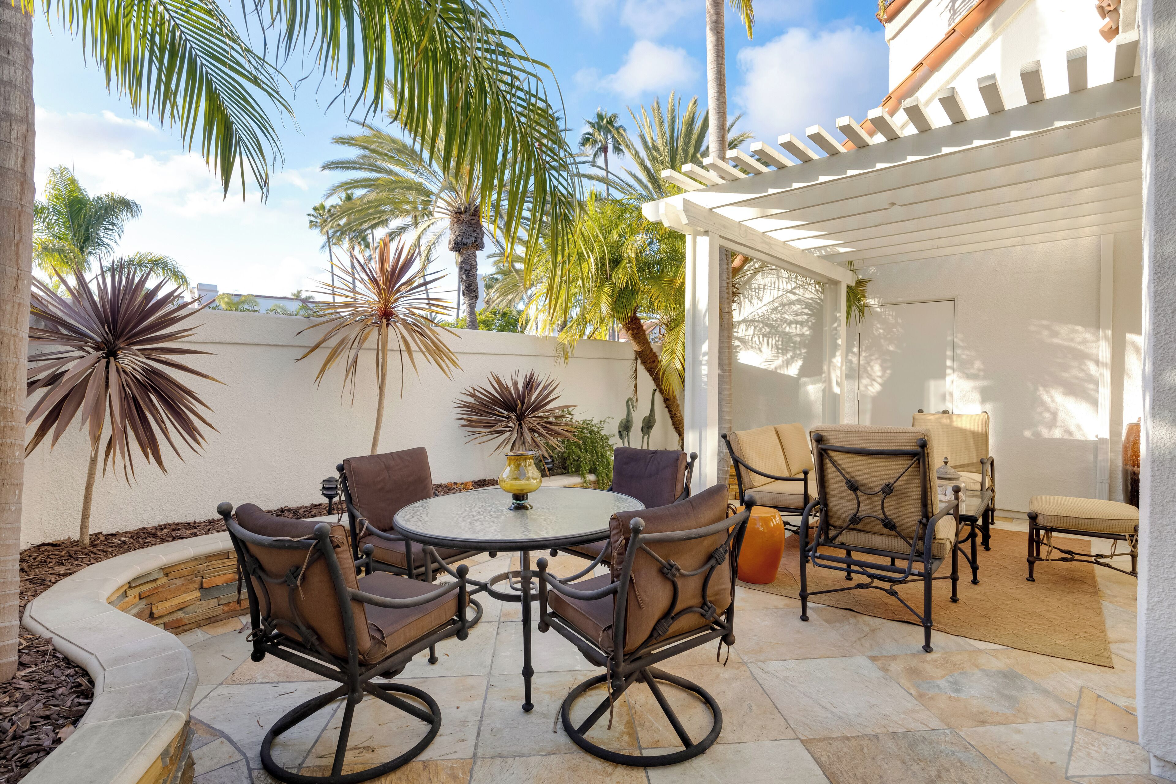 A patio area with a round table and chairs, surrounded by palm trees and a white pergola.