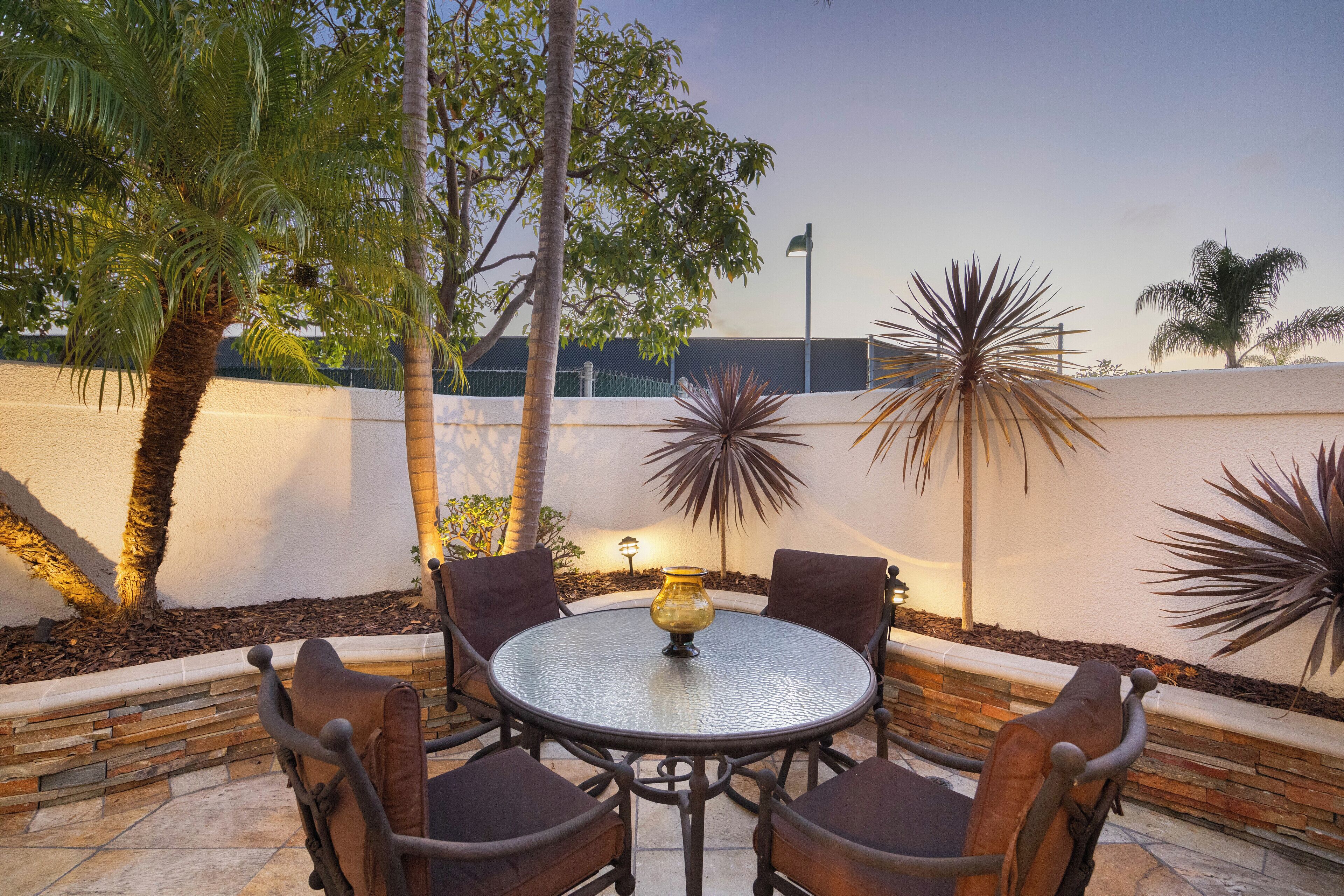 A round outdoor table with chairs, surrounded by a low stone wall, with palm trees and a fence in the background.