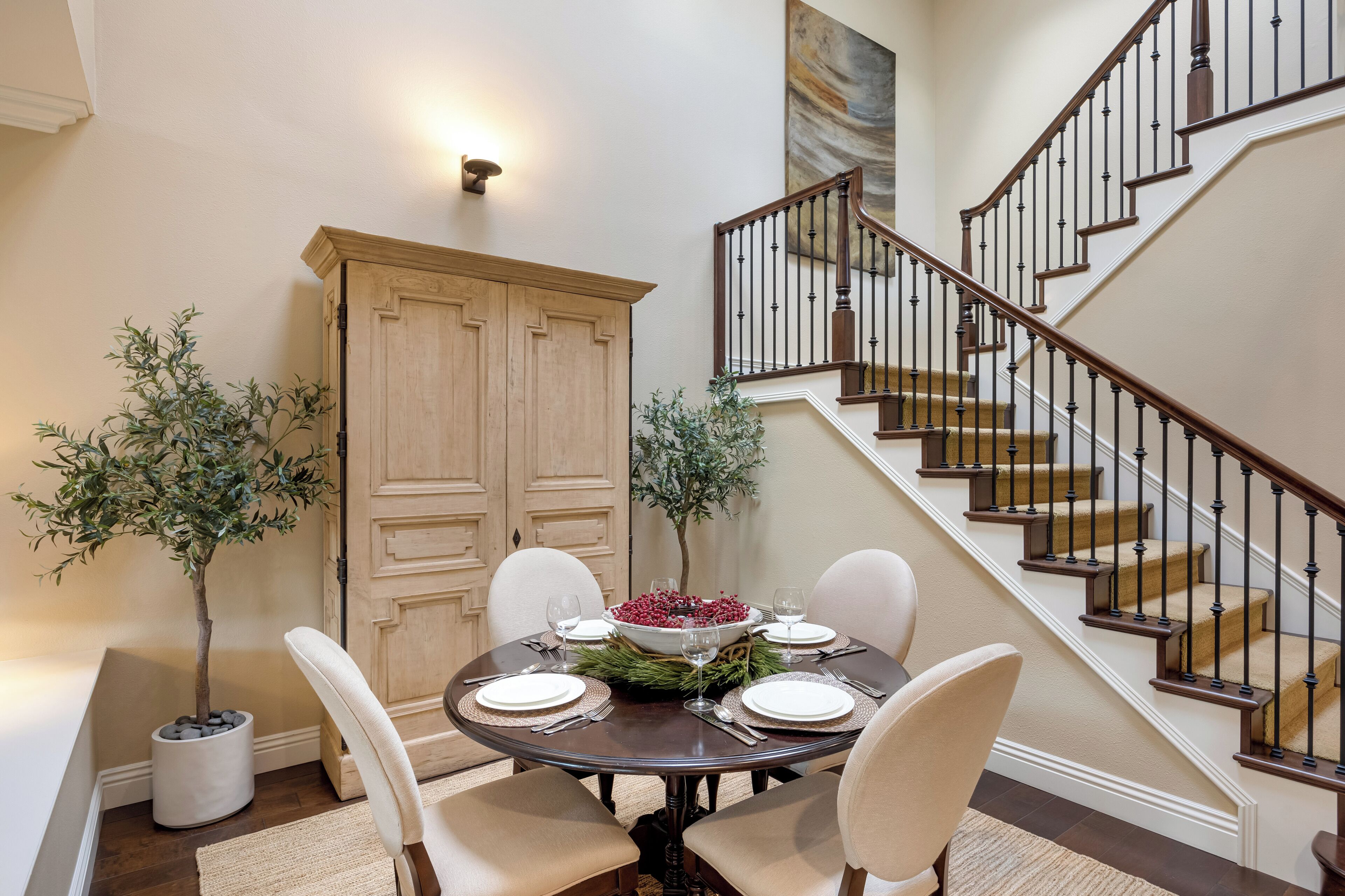 A dining area with a round table set for four, flanked by two potted plants, a wooden cabinet, and a staircase with a railing.