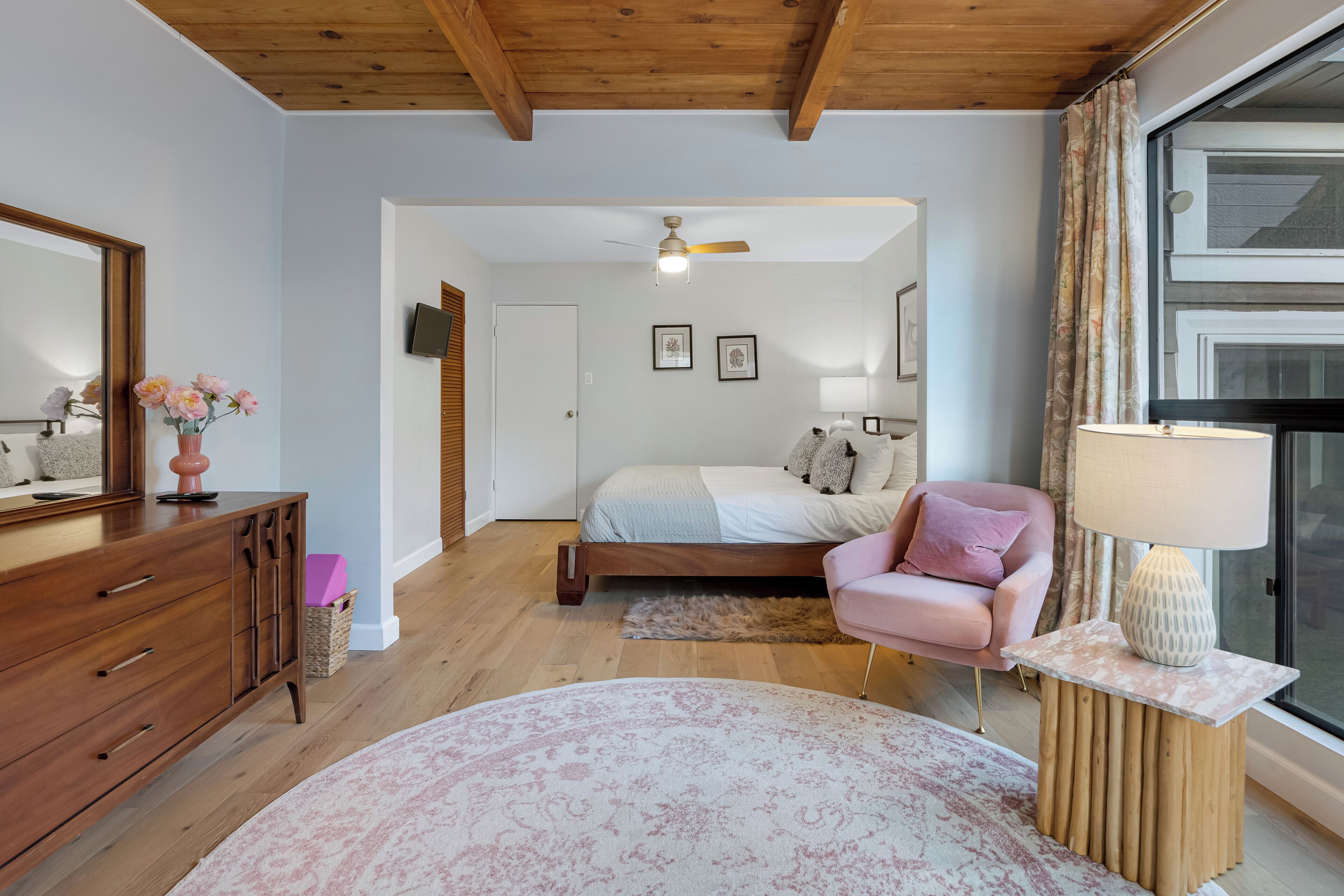 A bedroom with a wooden ceiling, a bed with a white and gray bedspread, a pink chair, a wooden nightstand with a lamp, and a large window with curtains.