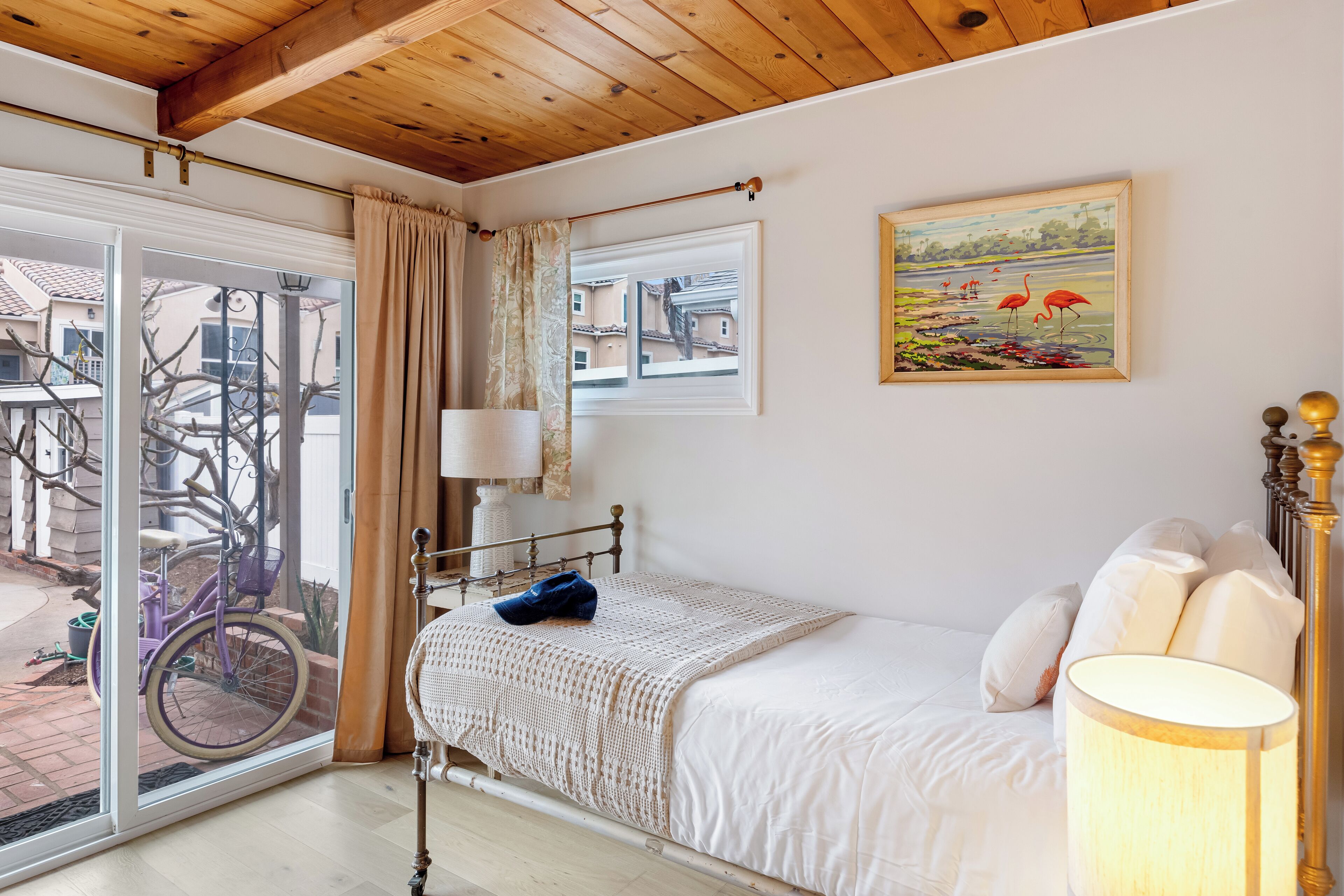 A bedroom with a bed, a lamp, a painting, and a bicycle outside the window.