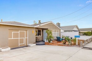 Une maison de plain-pied avec un garage, un patio couvert et un petit jardin agrémenté de plantes en pot et de meubles d’extérieur.