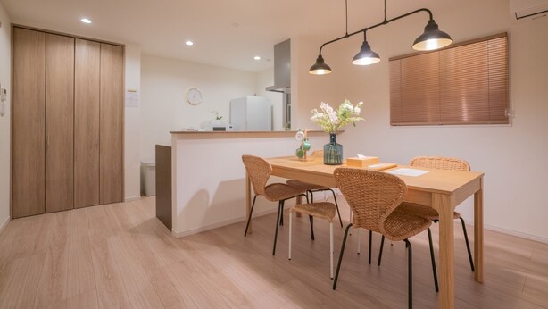 A dining area with a wooden table, four wicker chairs, and a vase of flowers. There is a kitchen in the background with a refrigerator and a sink.