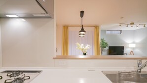 A modern kitchen with a gas stove, a sink, a window with curtains, a hanging pendant light, and a flat-screen TV mounted above the counter.