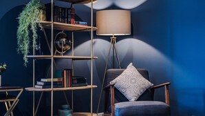 A modern living room with a dark blue wall, a wooden bookshelf, a floor lamp, and a blue armchair with a patterned pillow.