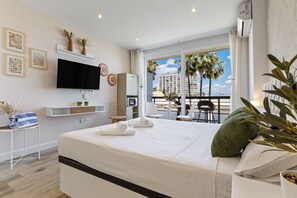 Ett hotellrum med en säng, TV, minibar och en balkong med utsikt över palmer och byggnader.