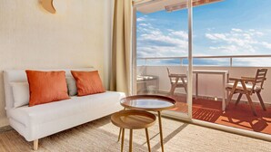 Habitación de hotel con un sofá blanco, dos almohadas naranjas, una mesita redonda pequeña y un gran ventanal corredero que da a un balcón con mobiliario exterior.