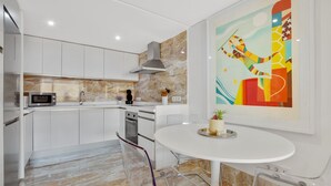 Una cocina moderna con armarios blancos, un microondas y una estufa. Hay una mesa de comedor con sillas y un cuadro abstracto y colorido en la pared.