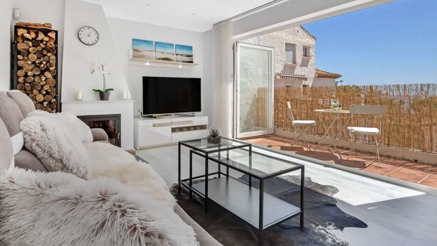 A modern living room with a large TV, a glass coffee table, and a sofa. There is a fireplace, a clock, and a view of a house through large glass doors. - Casa René - Pueblo Blanco (Torremolinos)