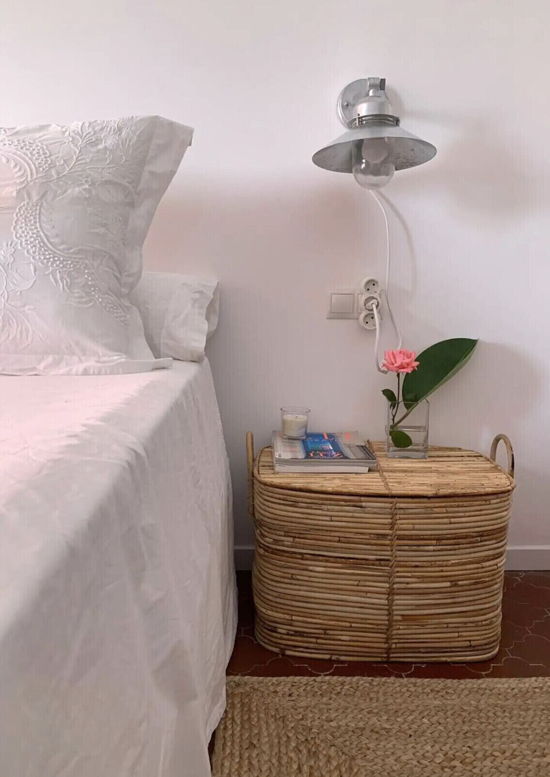 Une chambre à coucher avec un lit, une lampe de chevet, une table d’appoint en osier sur laquelle se trouvent un livre et une bougie, et une plante en pot.