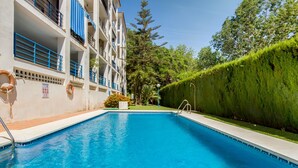 Une piscine à l’eau d’un bleu limpide, entourée d’un bâtiment blanc avec des balustrades bleues et une haie verte.