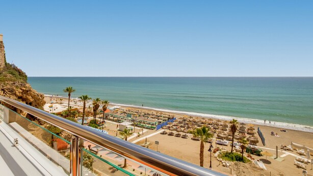 A beach view from a balcony with a clear sky, palm trees, and a sandy beach with umbrellas and lounge chairs.