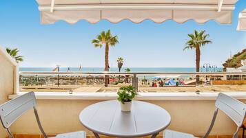 Un balcón con vistas a la playa, con una mesa y sillas, palmeras y cielo despejado.