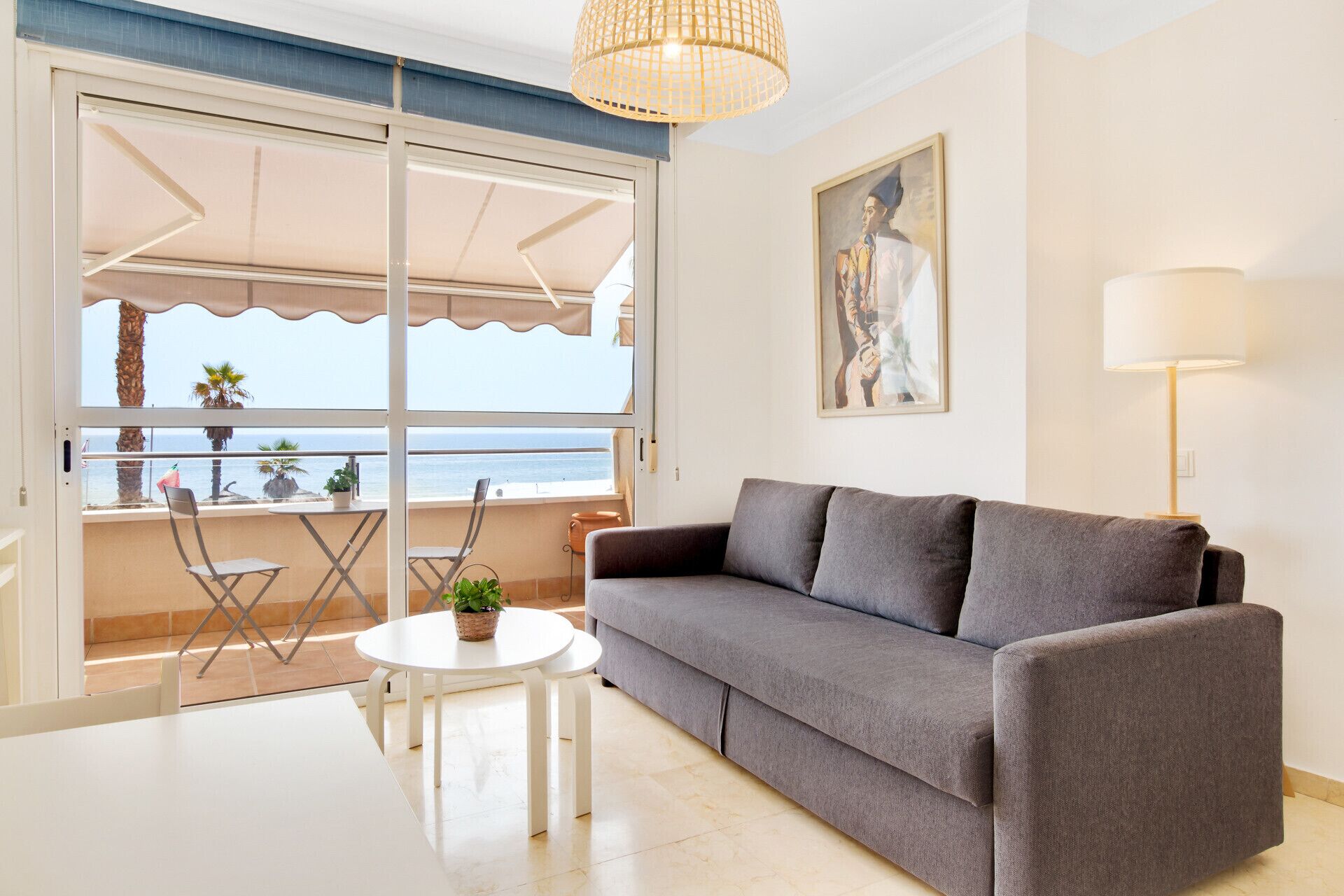 A living room with a grey sofa, a small white table, and a chair. There is a balcony with a view of the sea and a painting on the wall.