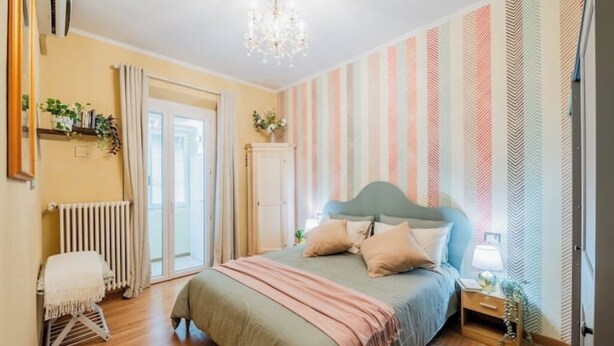 A bedroom with a bed, a radiator, a window with curtains, a chandelier, and a wall with striped wallpaper. - Aretina Apartment Cozy (Firenze)