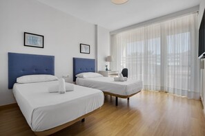 Two single beds with white linens and a rolled towel, a bedside table with a lamp, and a large window with sheer curtains.