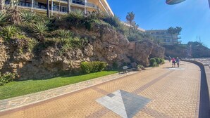 Eine Uferpromenade mit steilem Felsenhügel, Palmen und einem mehrstöckigen Gebäude mit Balkonen.