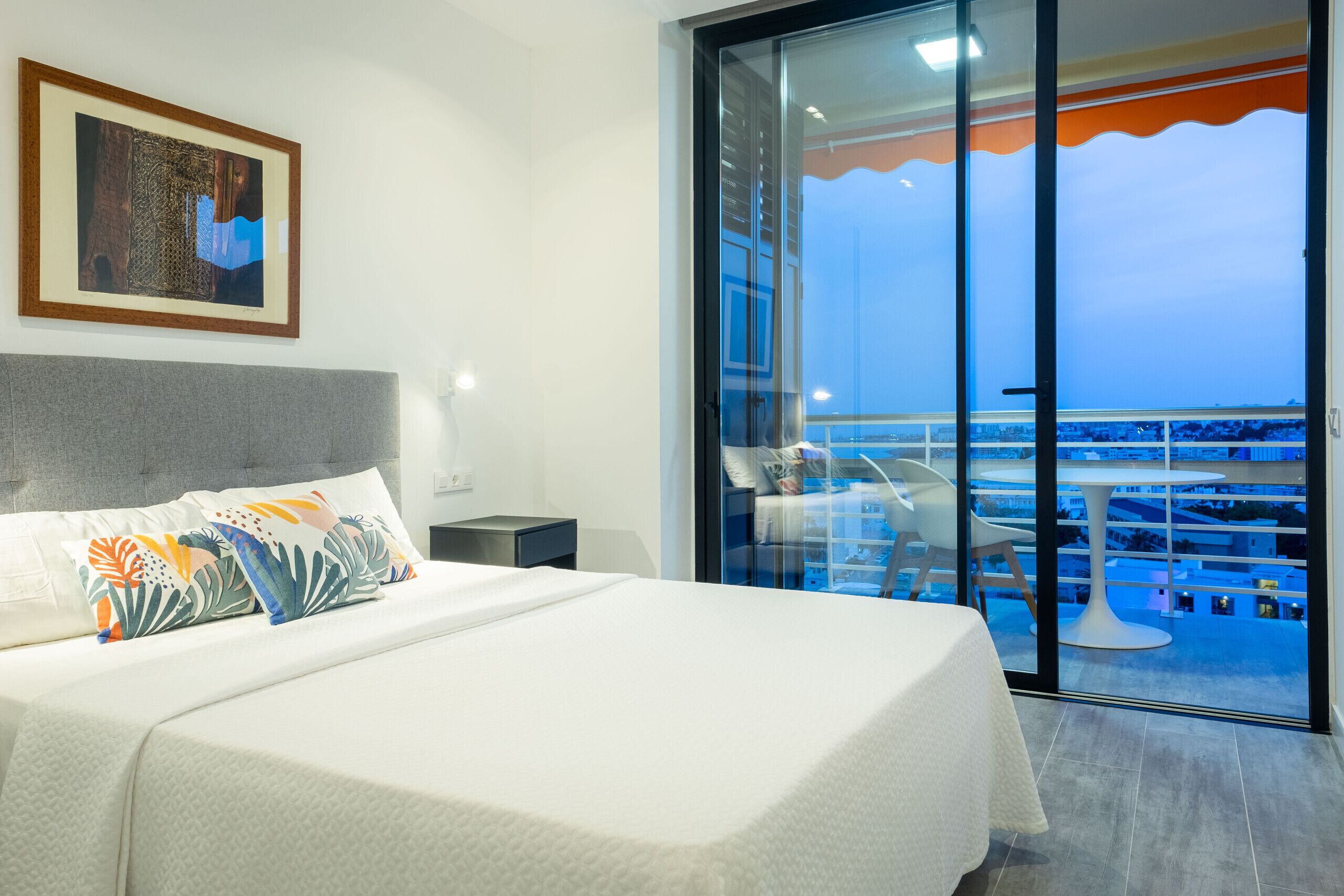 Habitación de hotel con una cama, una mesita de noche, un cuadro enmarcado y un balcón con vistas a la ciudad.