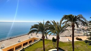 Vistes costaneres amb cel serè, palmeres, una platja i un edifici amb terrat pla que domina el mar.