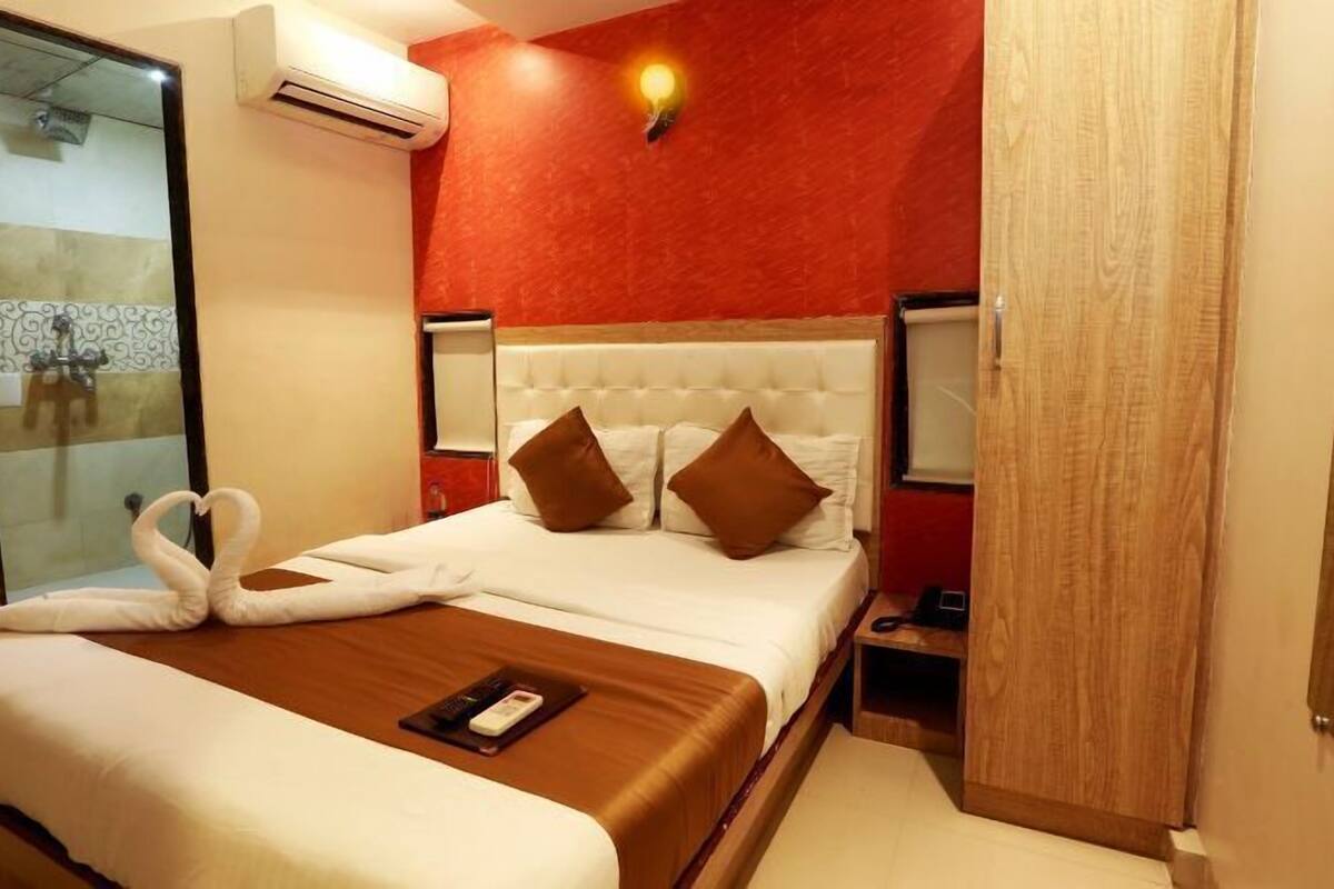 A hotel room with a bed, a bedside table, an air conditioning unit, a wall-mounted light, and a wooden door.
