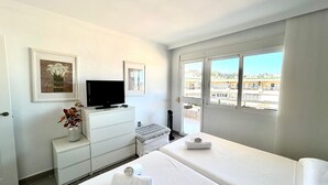 A hotel room with a bed, a TV on a dresser, a window with a view of buildings, and a small table with a vase of flowers. - Casa Hugo - La Carihuela (Torremolinos)